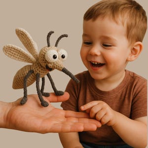 Puede incluir: Un juguete de mosquito de ganchillo beige y gris, con ojos grandes, alas y una larga probóscide. Un niño sonríe y señala el juguete. El juguete se sostiene en una mano abierta. Juguete hecho a mano.
