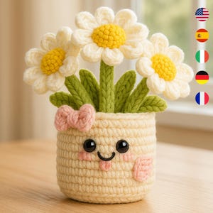 Peut inclure: Un pot de fleurs crocheté avec trois fleurs en forme de marguerites. Les fleurs ont des pétales blancs et des centres jaunes. Le pot est beige avec un visage souriant, des joues roses et un nœud. Les tiges et les feuilles vertes ajoutent une touche naturelle.