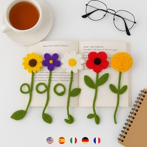 以下が含まれることがあります： 開いた本に、手作りのかぎ針編みの花のしおりが5つあります。各しおりは、ヒマワリ、紫の花、デイジー、赤いポピー、黄色のマリーゴールドなど、異なる花の模様が特徴です。しおりは、緑色の茎と葉が付いています。