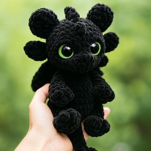 May include: A handmade black crochet dragon plushie with large green eyes. The dragon has a textured, rounded body, wings, and a tail. The plushie is held in a hand against a blurred green background.