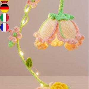 Puede incluir: Una lámpara de flores de ganchillo hecha a mano, con un tallo verde claro y una base crema. La lámpara presenta una gran pantalla en forma de flor en tonos amarillos y melocotón, con pequeñas luces LED. La base tiene flores de ganchillo rosas y amarillas.