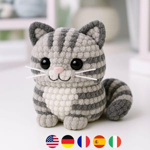 Może przedstawiać: Ręcznie robiona figurka kota na szydełku w odcieniach szarości i bieli. Kot ma duże czarne oczy, różowy nos i białe wąsy. Pasiasty wzór pokrywa ciało, ogon i głowę. Uroczy, miękki i przytulny przedmiot dekoracyjny.