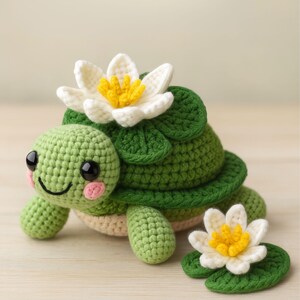 May include: A crocheted turtle, predominantly green, with a smiling face, pink cheeks, and black eyes. It has a white and yellow flower on its back and another on a green lily pad. The turtle's shell and limbs are detailed.