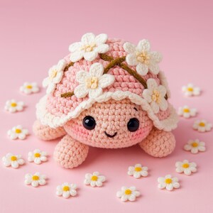 May include: A crocheted turtle with a pink shell decorated with white daisy flowers and a brown vine. The turtle has a smiling face with black eyes, surrounded by small daisy embellishments on a pink background.