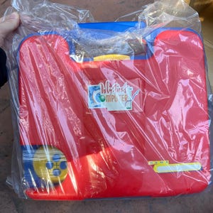 May include: A red and blue toy computer in a plastic wrapper. The toy has a handle and a yellow button. The text "My First Computer" is visible on the front. There is a space to write a name.