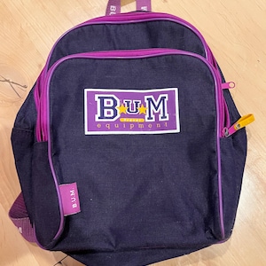 May include: A navy blue backpack with purple trim and accents. The front features a rectangular patch with the text "BUM equipment" in white and purple. The backpack has a top handle, adjustable straps, and a zippered front pocket.