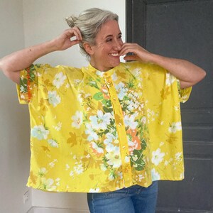 Pode incluir: Uma camisa amarela de botões com mangas curtas e estampa floral. A camisa tem um caimento solto e uma gola.