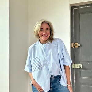 Pode incluir: Uma camisa azul claro de botões com um bolso xadrez vichy e mangas curtas. A camisa é usada com calças de ganga azuis.