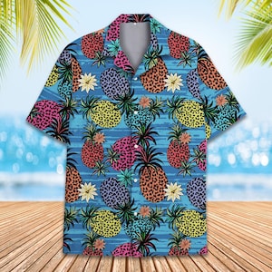 Könnte beinhalten: Kurzärmeliges Hemd mit einem lebendigen Ananasmuster. Das Hemd hat einen blauen Hintergrund mit horizontalen Streifen und farbenfrohen Ananas in Pink, Gelb, Lila und Orange. Es hat einen klassischen Kragen.