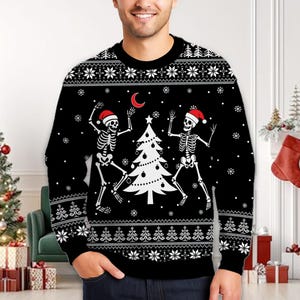 Puede incluir: Un suéter negro de manga larga con cuello redondo que presenta un gráfico blanco de dos esqueletos con gorros de Papá Noel rojos bailando alrededor de un árbol de Navidad blanco. El diseño incluye una luna creciente roja, copos de nieve y un borde geométrico.