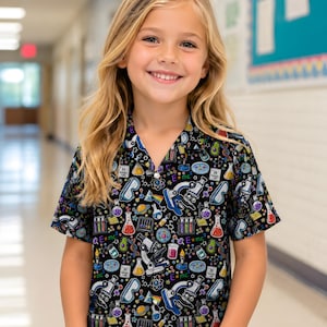 May include: A short-sleeved button-down shirt with a black background featuring a colorful science-themed print. The pattern includes illustrations of microscopes, test tubes, beakers, safety goggles, atoms, and the text E=mc2 in various colors.