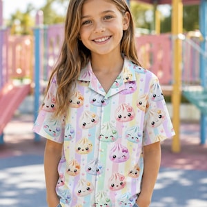 May include: A short-sleeved button-down shirt featuring a pastel vertical striped pattern and a repeating print of cute, smiling dumpling characters with small decorative accents like bows and flowers in shades of pink, blue, yellow, and green.