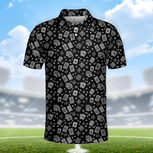 May include: Black polo shirt with a repeating pattern of playing cards in white. The shirt has a collared neckline and short sleeves. The background is a blurred image of a stadium and a blue sky.