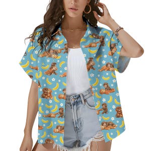 Può includere: Camicia azzurra con bottoni e un motivo giocoso di scimmie marroni, banane gialle e fiori bianchi. La camicia a maniche corte è realizzata in un tessuto leggero, perfetta per un look tropicale casual.