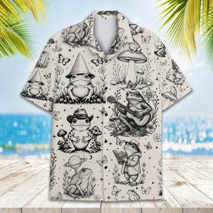 May include: White button-down shirt with a whimsical frog-themed pattern. The design features various frog illustrations, mushrooms, butterflies, and floral elements in a black and white sketch style. The shirt has short sleeves and a classic collar.