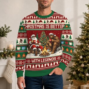 May include: A long-sleeve crewneck sweatshirt in red, green, and white featuring a graphic of Santa Claus and Bigfoot sitting by a campfire. The text CHRISTMAS IS BETTER WITH LEGENDS is printed across the front, surrounded by patterns of trees, mugs, and footprints.