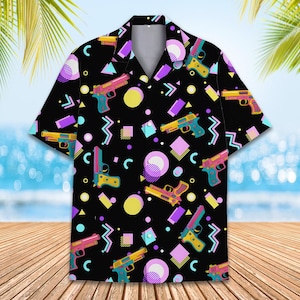 Puede incluir: Una camisa negra de botones con un patrón geométrico colorido que presenta imágenes estilizadas de pistolas. El patrón incluye colores rosa, azul, amarillo y verde.