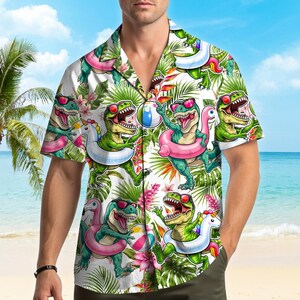 May include: A short-sleeve button-down shirt featuring a white background with a repeating pattern of green dinosaurs wearing pink sunglasses. The dinosaurs are depicted with colorful pool floats, beach balls, and tropical palm leaves and flowers.