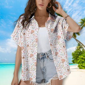 Könnte beinhalten: Weißes Button-Down-Hemd mit Blumen- und Ballerina-Print. Das Hemd hat kurze Ärmel und eine lockere Passform. Das Muster umfasst rosa Blumen, grüne Blätter und Balletttänzerinnen. Das Hemd wird mit Jeansshorts kombiniert.