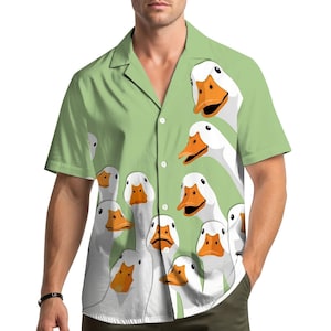 May include: A light green short-sleeve button-down shirt featuring a repeating graphic print of white ducks with orange beaks. The shirt has a notched collar, a front button placket with white buttons, and a straight hemline across the bottom.