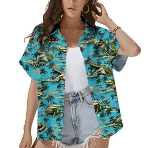 May include: A short-sleeved, button-down shirt with a turquoise background featuring a repeating pattern of green alligators, palm trees, and orange suns. The shirt is open, revealing a white tank top and denim shorts.