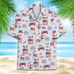 May include: White button-up shirt with a patriotic pattern featuring red, white, and blue American flags, fireworks, and flamingos wearing party hats. The shirt has a repeating pattern of "Happy 4th of July" text.