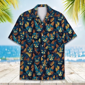 May include: Short-sleeved button-down shirt featuring a repeating pattern of stylized cat faces in shades of blue, orange, and brown. The shirt has a classic collar and a relaxed fit, perfect for casual wear. The background shows a beach scene.