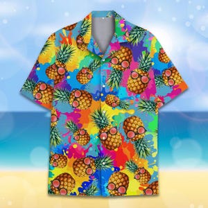 May include: Short-sleeved button-down shirt with a colourful design. The shirt displays a pattern of pineapples wearing sunglasses against a backdrop of bright paint splatters in various colours, including blue, yellow, pink, and green.