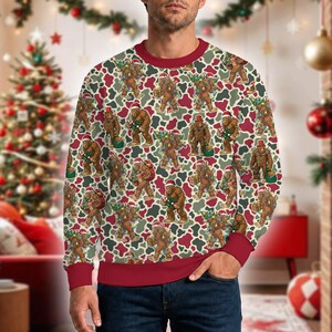 Peut inclure: Un sweat-shirt à col rond et manches longues avec un motif répétitif de Bigfoot marron portant des bonnets de Père Noël, transportant des sapins et des cadeaux sur un fond camouflage aux accents rouges, blancs et verts.
