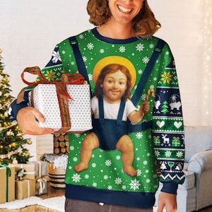 May include: A green long-sleeve sweatshirt featuring a graphic of a person with a halo in a baby carrier, surrounded by white snowflakes. The sleeves have dark blue cuffs and a pattern of reindeer, trees, and gifts. A gift box is held in front.