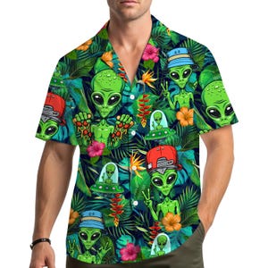 May include: A short-sleeved button-down shirt featuring a vibrant pattern of green aliens, UFOs, and tropical flowers set against a dark blue background of lush foliage. The aliens are depicted with various accessories, including hats. The shirt has a relaxed, casual style.