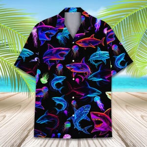 May include: Black short-sleeved shirt with a colourful pattern of neon sharks and jellyfish. The design features shades of blue, pink, purple, and orange, contrasting against the black background. The shirt has a classic collar and button-up front.