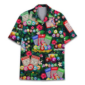 May include: Short-sleeved shirt with a bingo-themed design. The shirt displays bingo cards, colourful bingo balls, and the word "BINGO" in multiple locations, set against a dark green tropical leaf background.