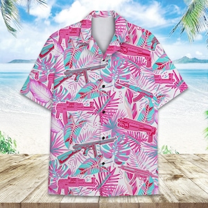 Puede incluir: Camisa de manga corta con estampado tropical. La camisa presenta un fondo blanco con un diseño repetido de armas rosas y hojas tropicales turquesas y rosas. La camisa se muestra sobre una superficie de madera con una escena de playa al fondo.