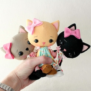 以下が含まれることがあります： ピンクのリボンと刺繍された顔をしたフェルトの猫のおもちゃ3つ。猫は茶色、オレンジ、黒です。