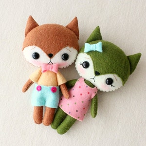May include: Two felt fox plushies, one brown with a blue bow tie and blue pants, the other green with a pink dress and a blue bow.
