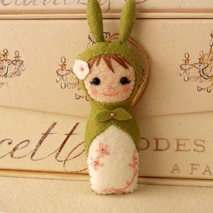 May include: A green felt bunny doll with a white felt dress and a pink flower on its head. The doll has brown hair and black eyes. The doll is sitting on a white surface.