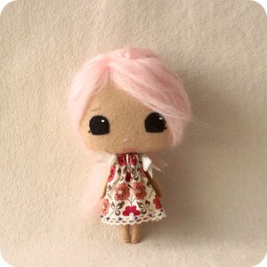 May include: A handmade doll with pink hair, large black eyes, and a floral dress. The doll is made of fabric and has a sweet, whimsical expression.