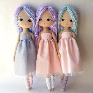 May include: Three handmade dolls with long, colorful yarn hair. Each doll is wearing a different colored dress. The doll on the left is wearing a gray and white striped dress, the doll in the middle is wearing a pink dress, and the doll on the right is wearing a light pink dress.