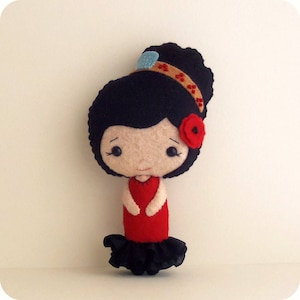 May include: A felt doll with black hair, a red dress, and a black ruffle skirt. The doll has a red flower on its head and a brown headband with blue accents.