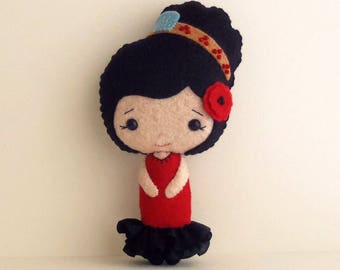 May include: A felt doll with black hair, a red dress, and a black ruffle skirt. The doll has a red flower on its head and a brown headband with blue accents.
