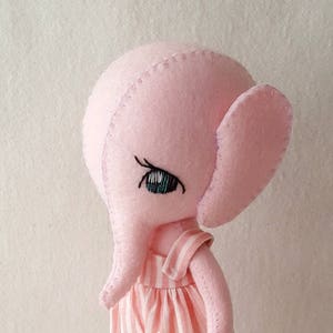 May include: A pink felt elephant doll wearing a pink and white striped overall with a floral patch. The doll has embroidered eyes and a stitched nose.