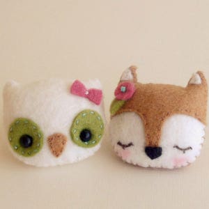 May include: A white felt owl with green eyes and a pink bow, and a brown felt fox with closed eyes and a pink flower.