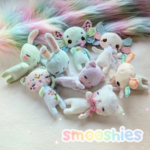 Op de afbeelding: Een verzameling handgemaakte pluche speelgoed in verschillende pastelkleuren, waaronder konijnen, katten en zeedieren. Elk pluche heeft geborduurde details en een grillig ontwerp. Het woord "Smooshies" is in een kleurrijk lettertype geschreven.