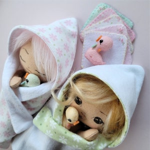 May include: Two handmade cloth dolls with blonde and pink hair are dressed in white and green fabric with floral patterns. Each doll is holding a small stuffed bird.