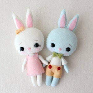 May include: Two felt bunny dolls, one white with a pink polka dot dress and a yellow bow, the other light blue with a yellow bow tie and orange shorts.