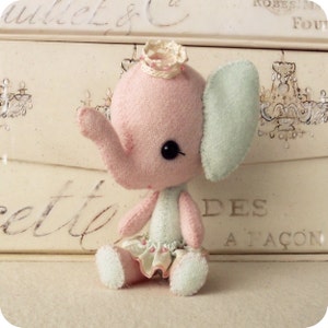 Könnte beinhalten: Ein rosa und blaues Filz-Elefantenspielzeug, das eine weiße Krone und einen rosa-weißen Rüschenrock trägt. Das Spielzeug sitzt auf einer weißen Oberfläche mit einer dekorativen Schachtel im Hintergrund.