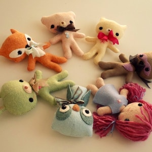 May include: A group of handmade felt animal toys, including a fox, a bunny, a bear, a cat, a bird, and a girl with pink hair. The toys are all different colors and have embroidered details.