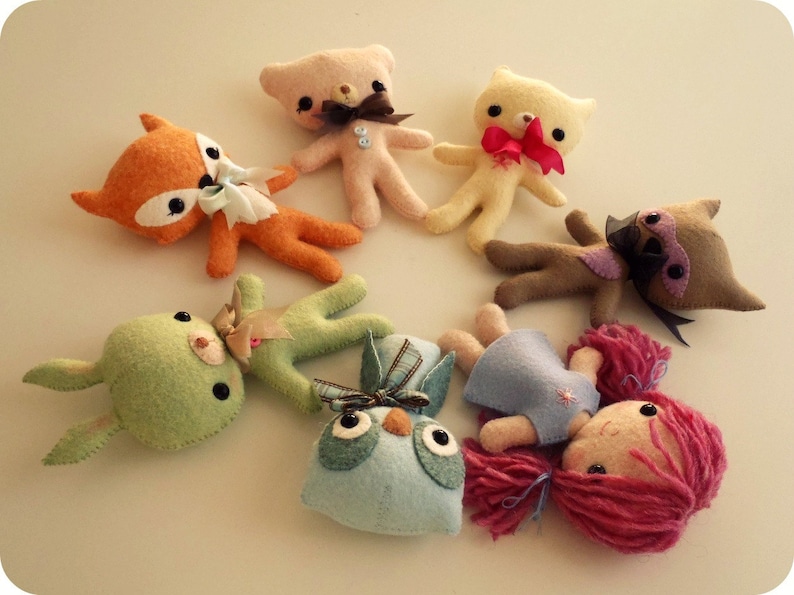 May include: A group of handmade felt animal toys, including a fox, a bunny, a bear, a cat, a bird, and a girl with pink hair. The toys are all different colors and have embroidered details.
