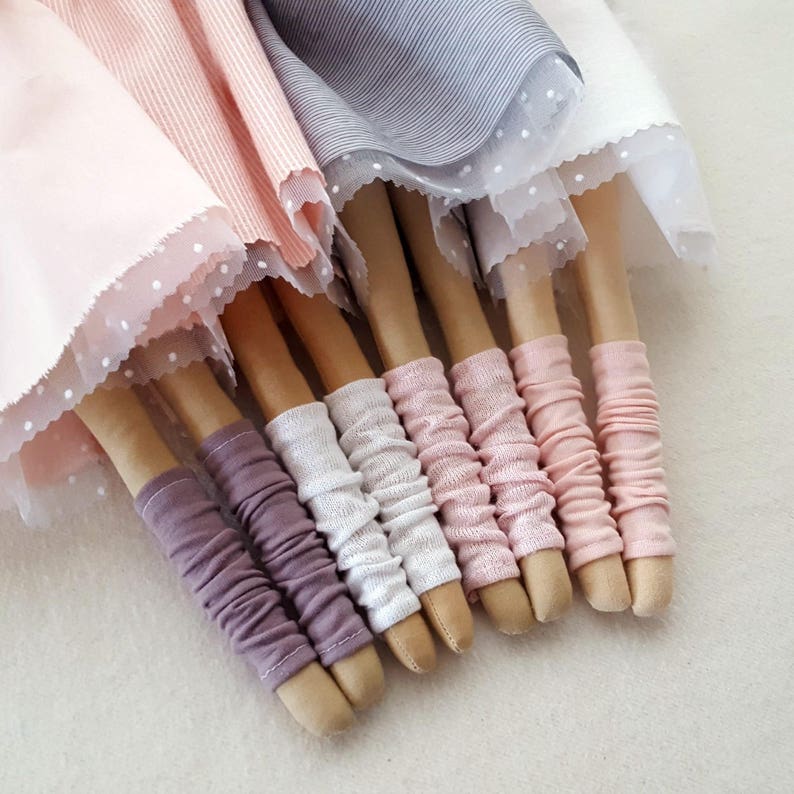 May include: A group of six wooden dolls with fabric skirts and leggings. The dolls are wearing pink, white, and purple leggings. The skirts are made of pink, gray, and white fabric.